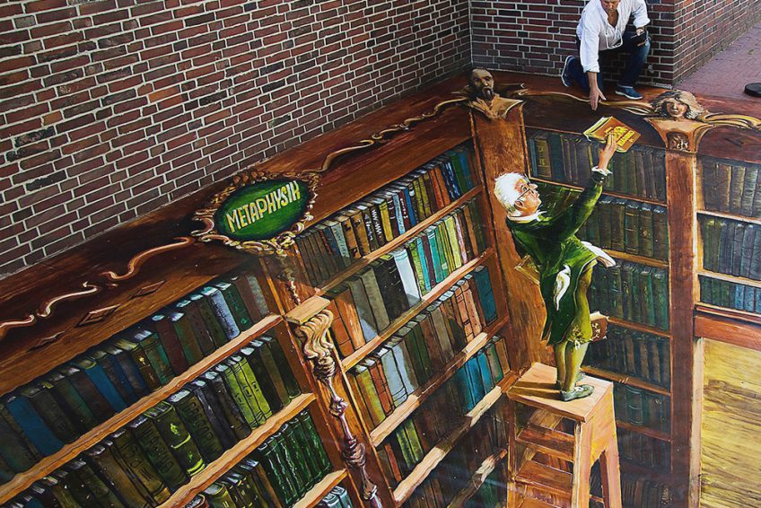 A person stands beside a pavement painting of towering library shelves, appearing to reach into the painted space.